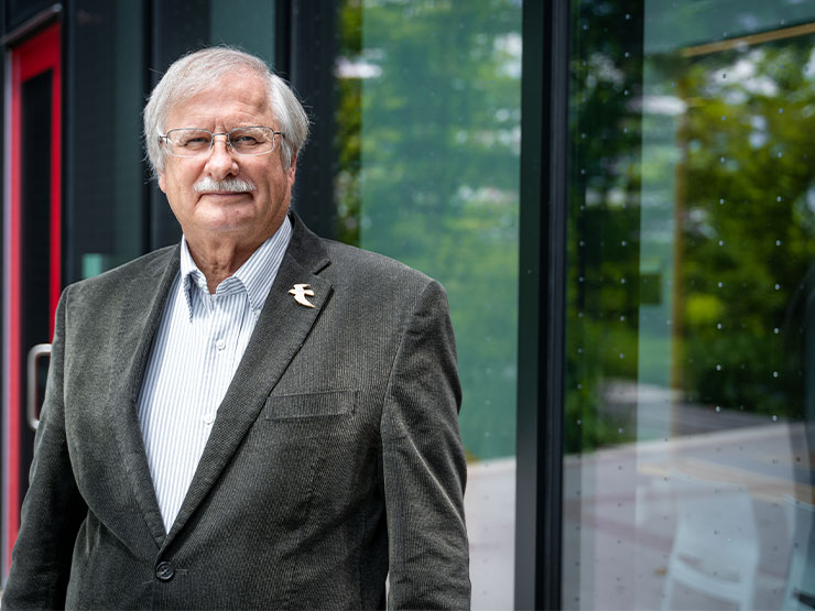 A man in a suit jacket stands outdoors near windows imprinted with faint dots to help deter bird collisions.