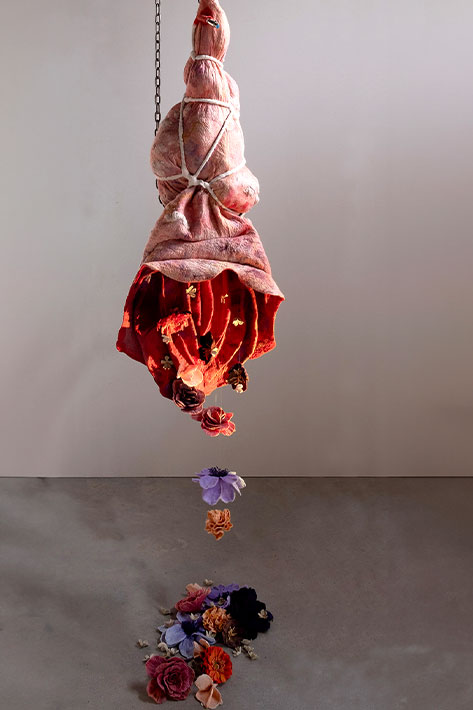 An artwork installment made of colorful fiber that appears to show a bouquet hanging upside down with blossoms falling to the ground beneath.