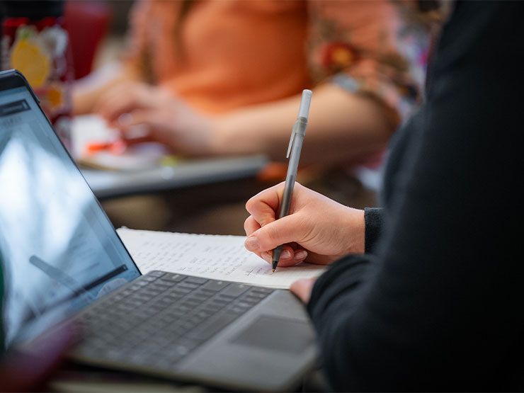 A student writes on a notebook next to an open laptop in a classroom.