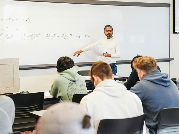 Economics professor Ranajoy Ray-Chaudhuri points to an equation on a whiteboard in front of a classroom.