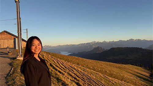 Fuka Aizawa stands smiling beside a field on a scenic vista in Switzerland.