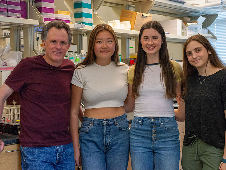 A college professor stands beside a group of college students in a biology lab.