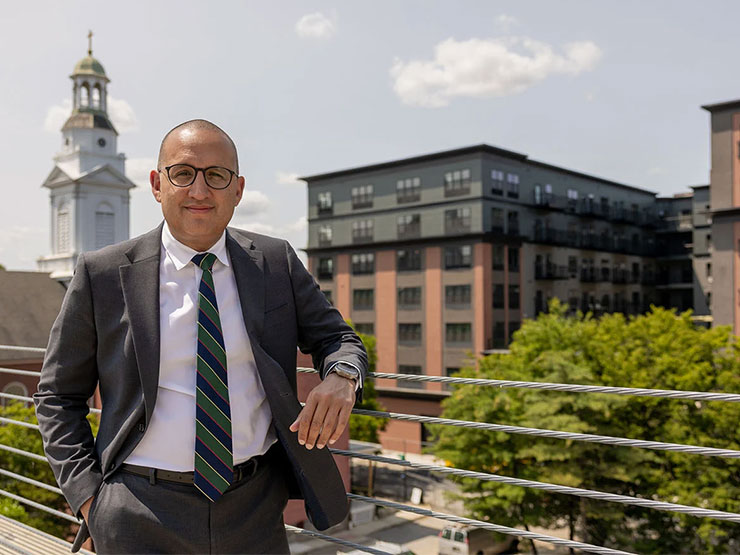 A man in a suit and glasses poses against a railing overlooking a city.