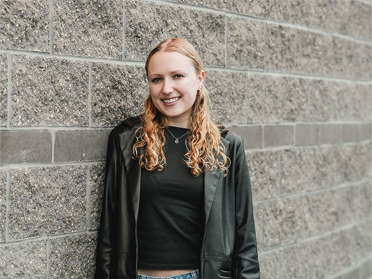 Daisy Cunningham, smiling, stands against a grey brick wall.