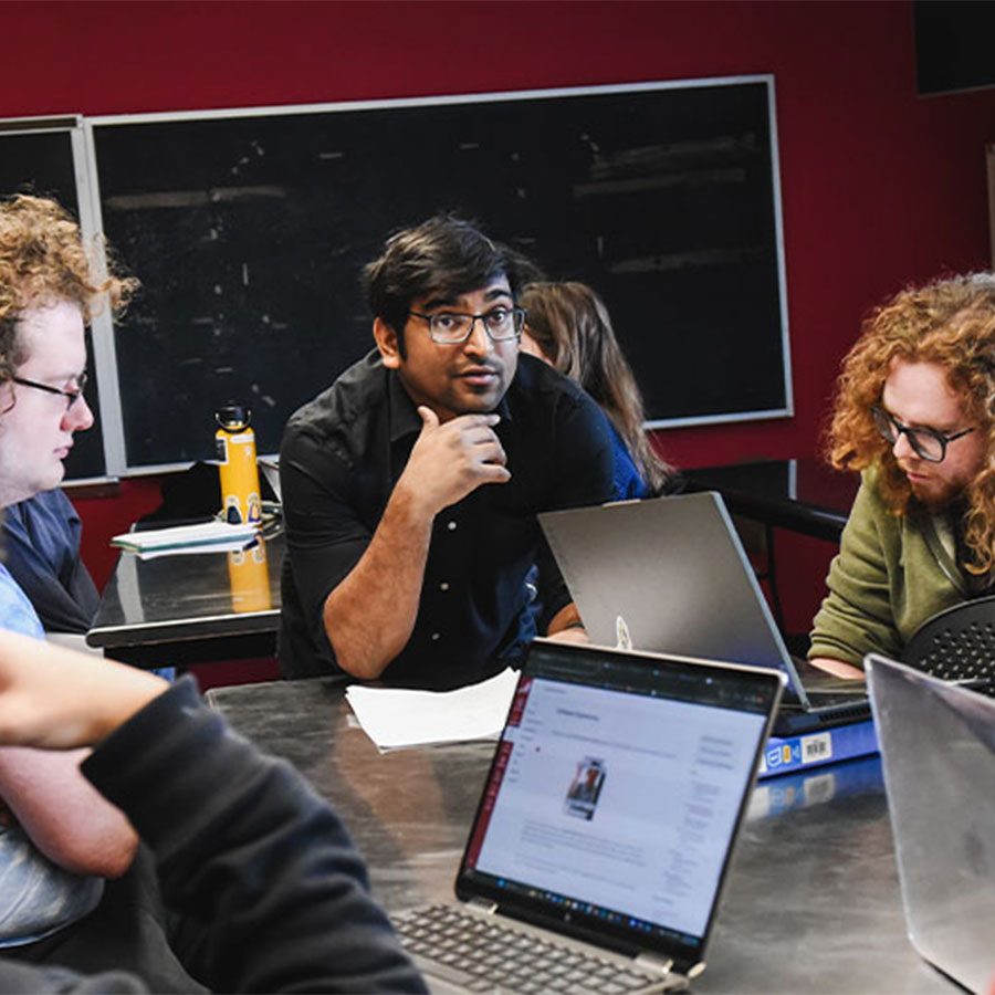 Proyash Podder leans over a table and speaks with a group of students working on open laptops.