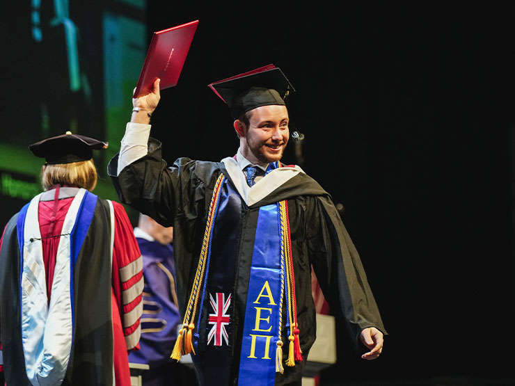 A recent Muhlenberg College grad grins while watching across the stage during commencement, holding a diploma overhead.