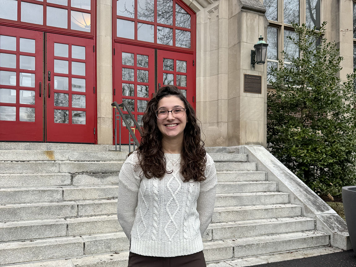 A college student in a gray sweater smiles in front of red doors