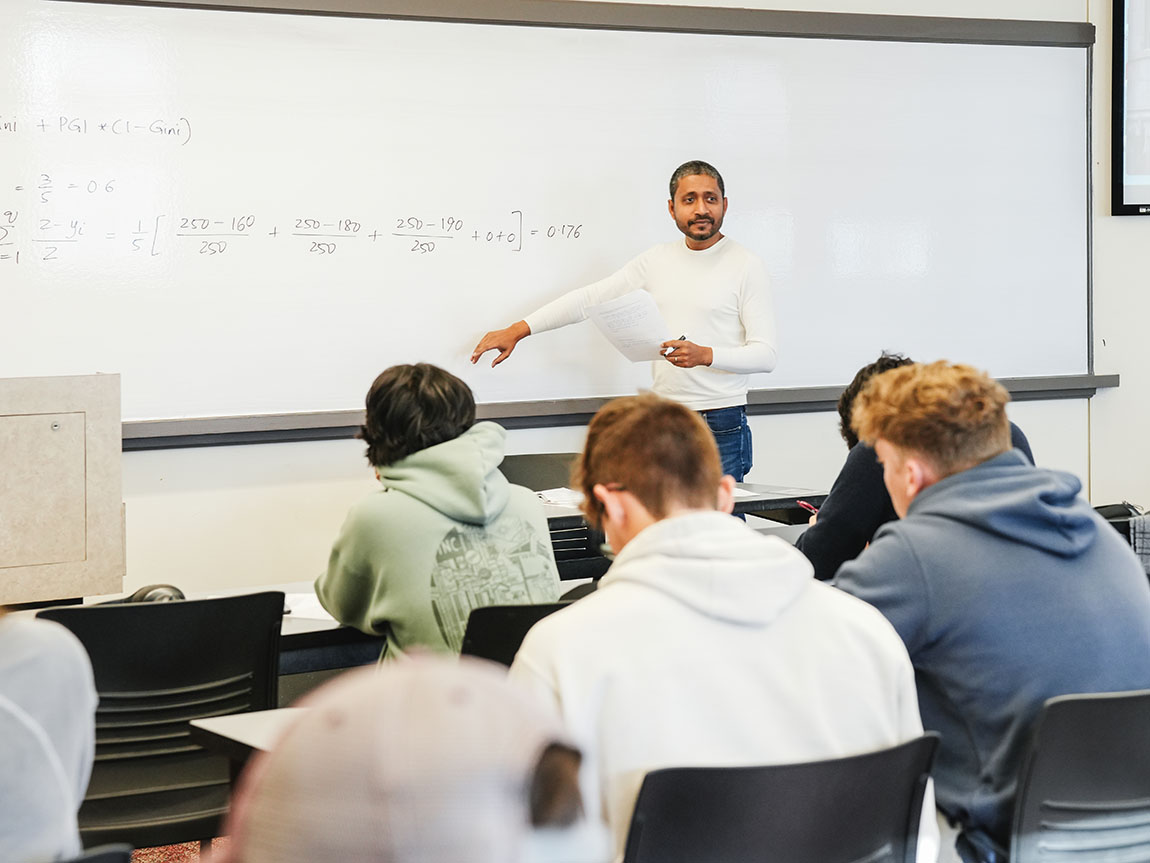 A college professor stands in front of a classroom and points at the whiteboard
