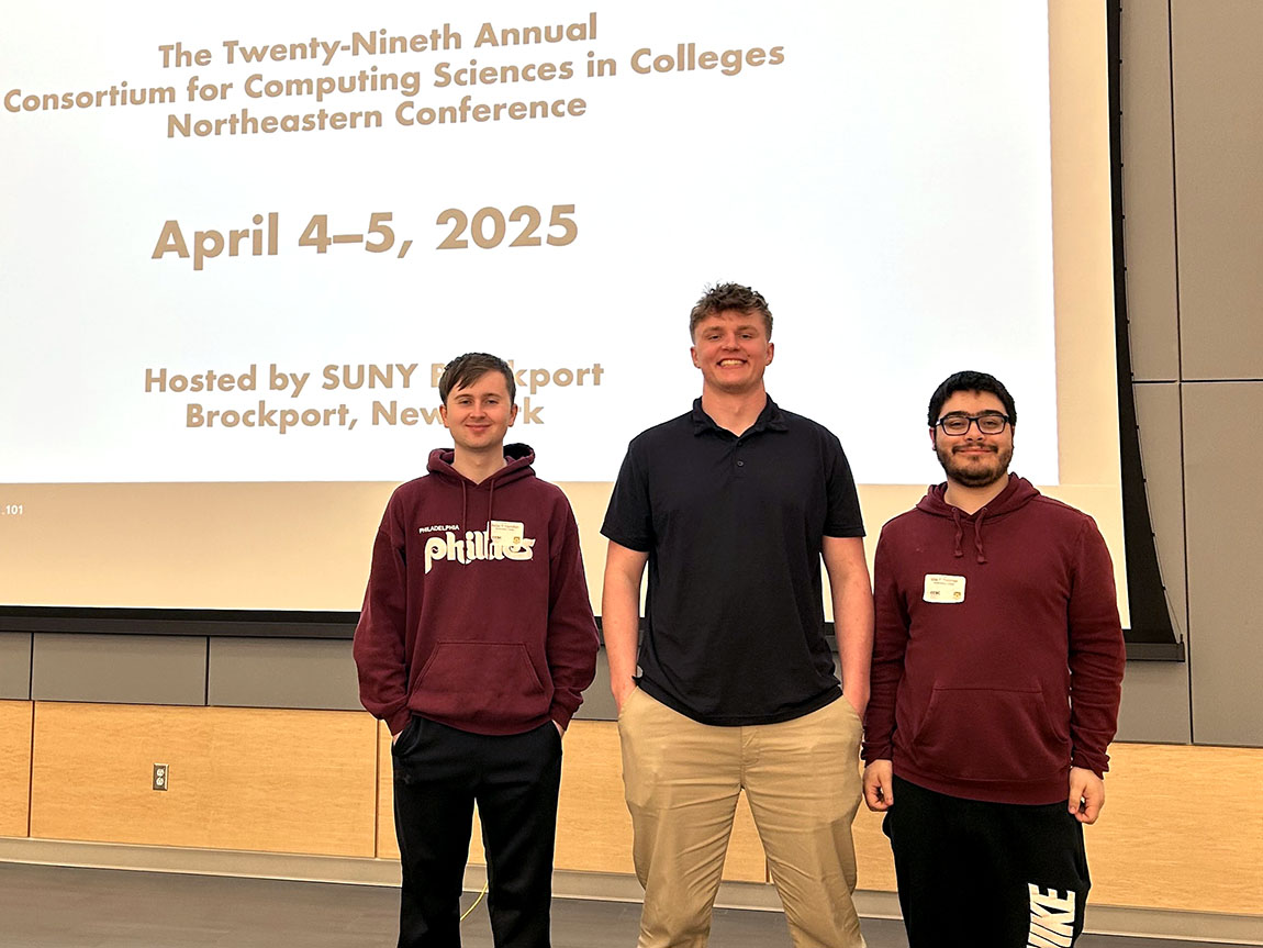 Three college students stand in front of a screen with a projection that reads the Twenty-Ninth Annual Consortium for Computing Sciences in Colleges Northeastern Conference