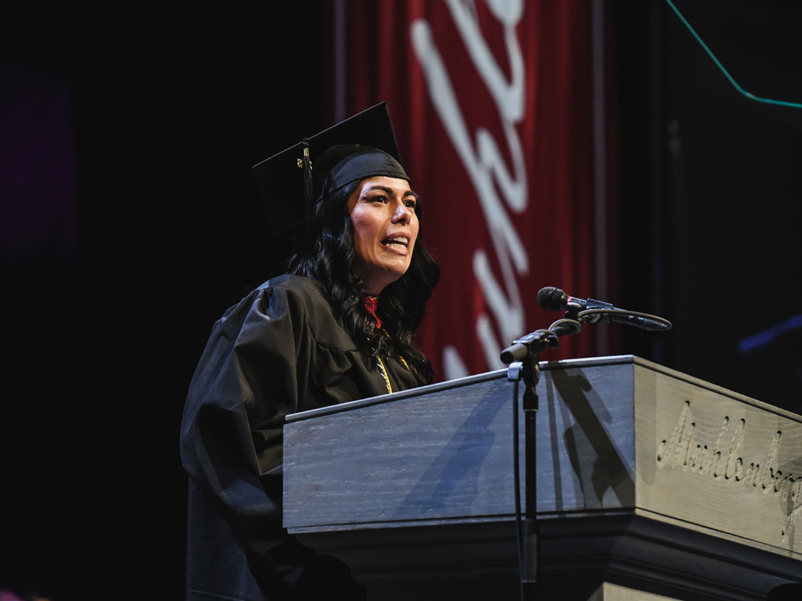 An adult student in a cap and gown stands at a podium and gives an address