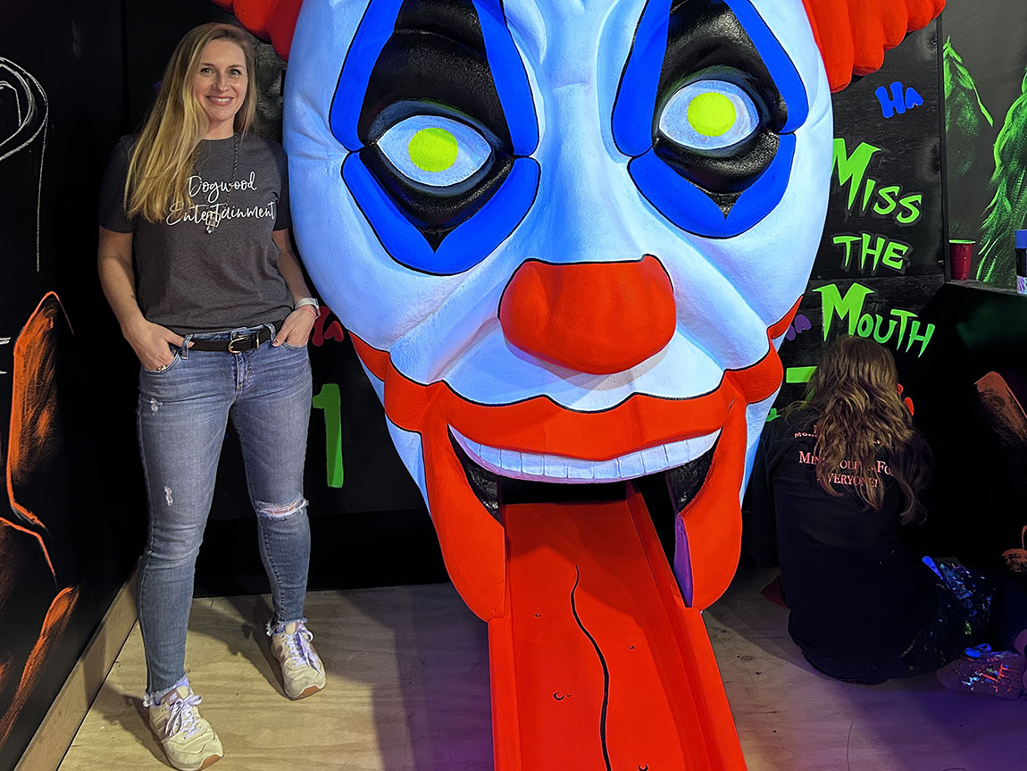 A woman wearing a shirt that says Dogwood Entertainment stands next to a giant evil clown head