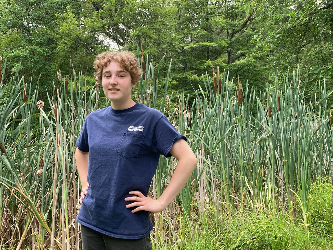 A college student in a dark blue T-shirt stands in front of high grass