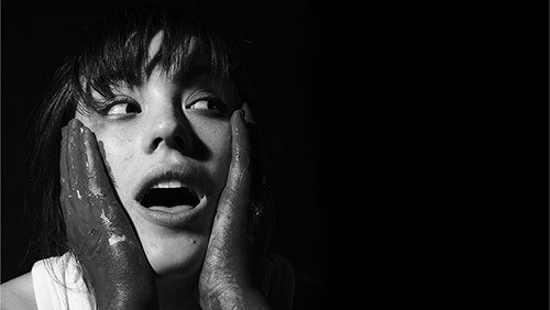 An person holds painted hands to face with a surprised expression in stylized black and white.