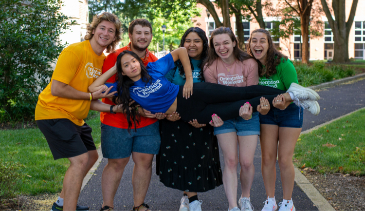 A group of students holding up another student outdoors on a college campus.